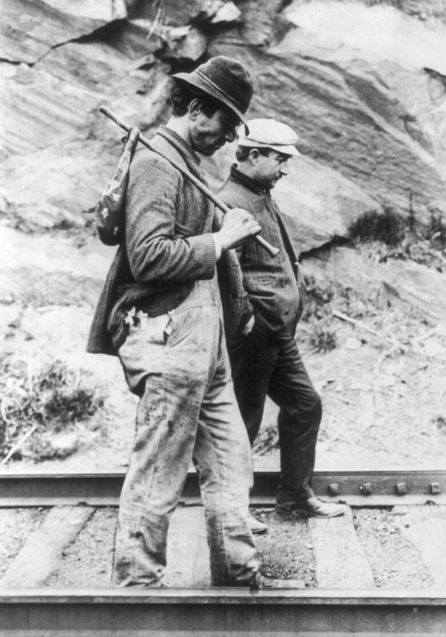 Two hobos walking along railroad tracks, one carrying a bindle